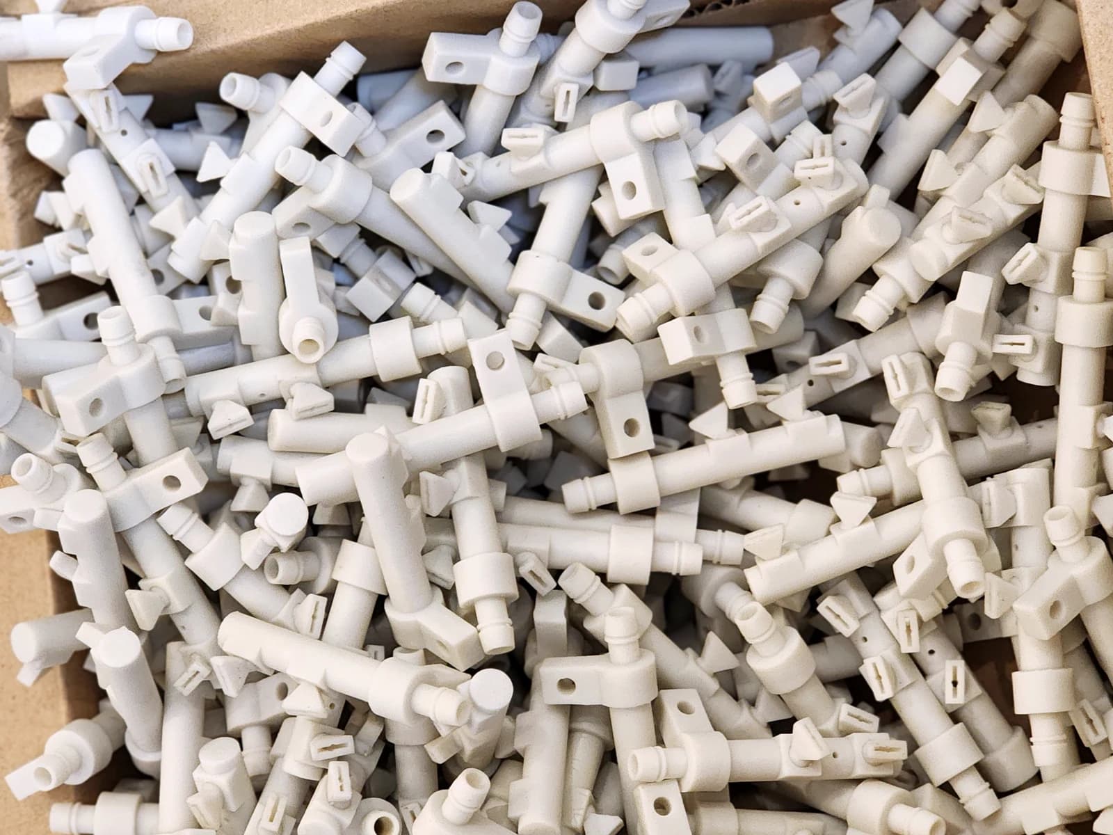 A batch of white tubular parts with mounting holes and tabs in a cardboard box.