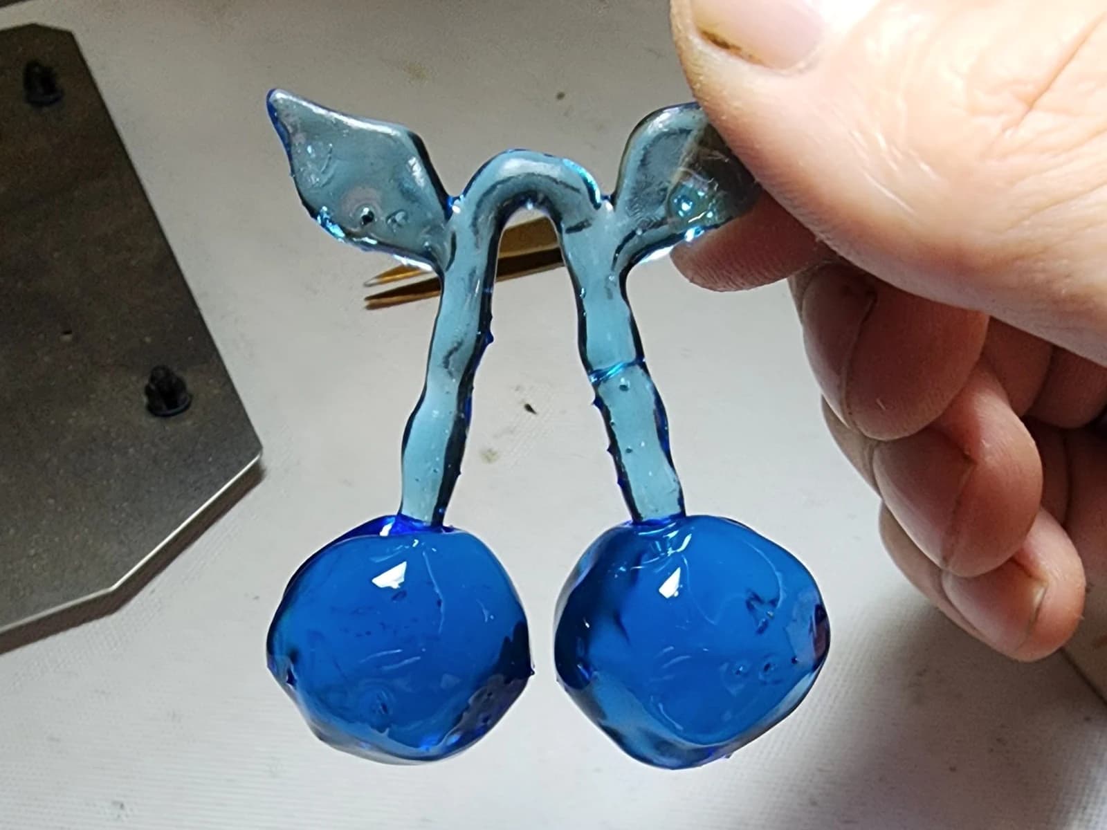 A translucent blue pendant shaped like two cherries with stems and leaves, held in a hand.