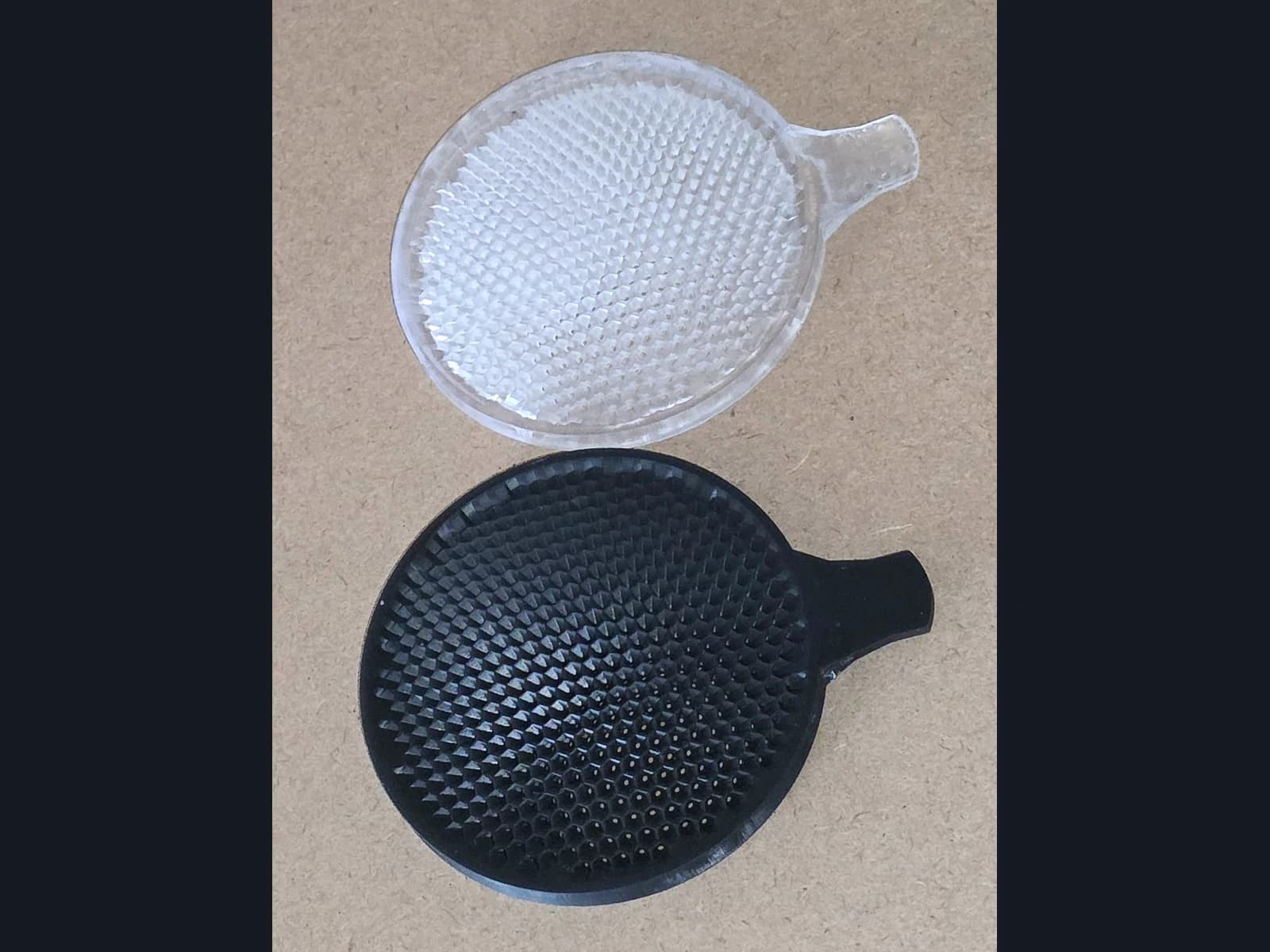Pale and black circular mesh parts with side tabs on a beige surface.