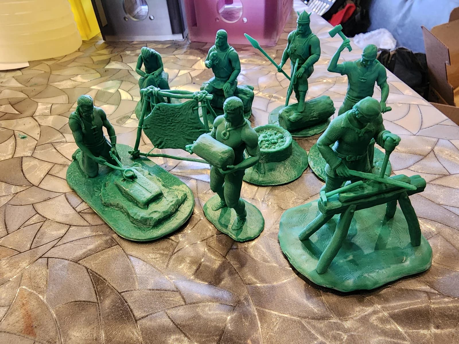 Green miniatures of people with tools and workpieces, including figures at an anvil and a workbench, on textured bases.
