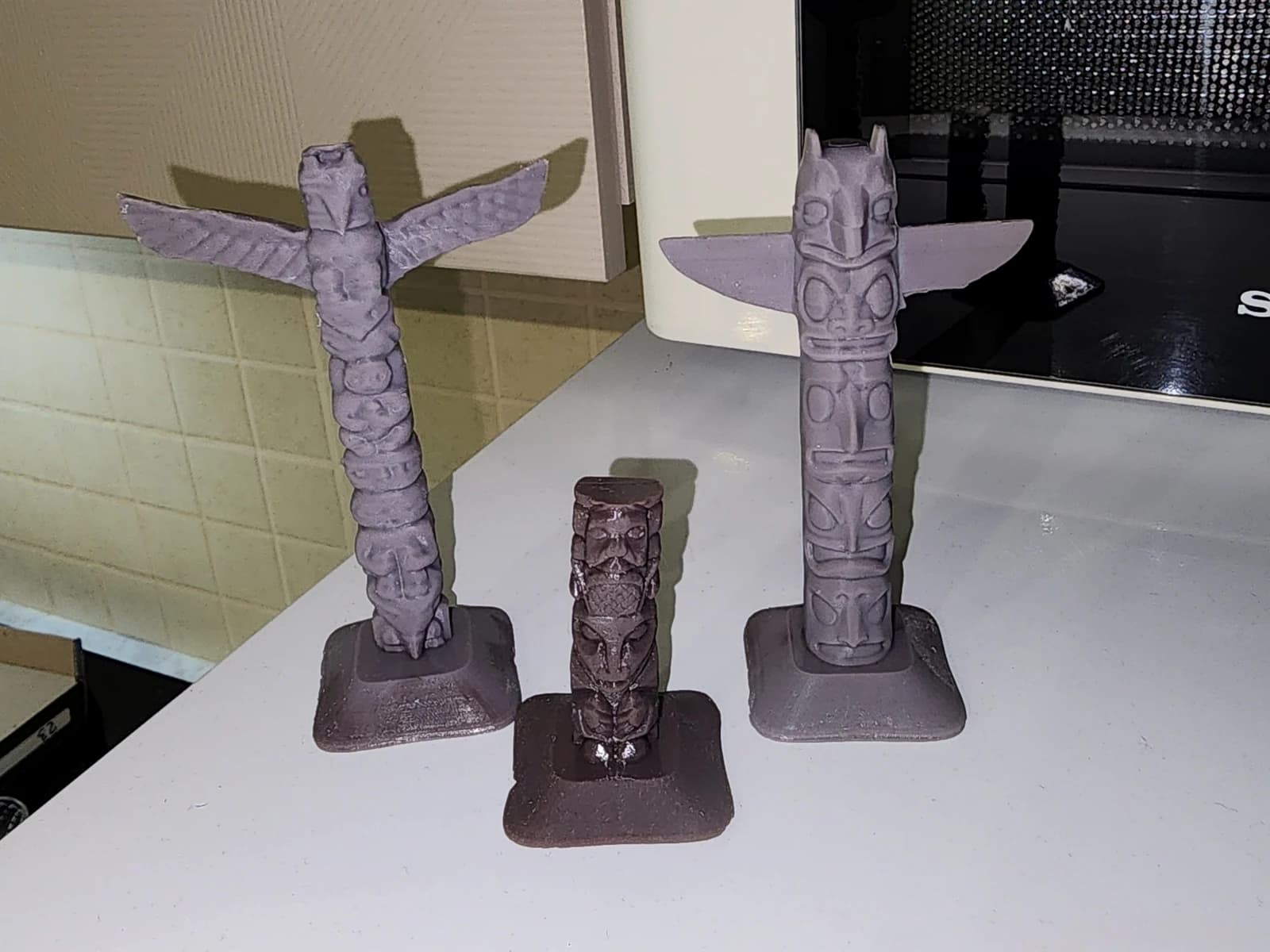 Three decorative poles on rectangular bases: two tall pieces with outstretched wings and one shorter piece, with raised faces along their surfaces.