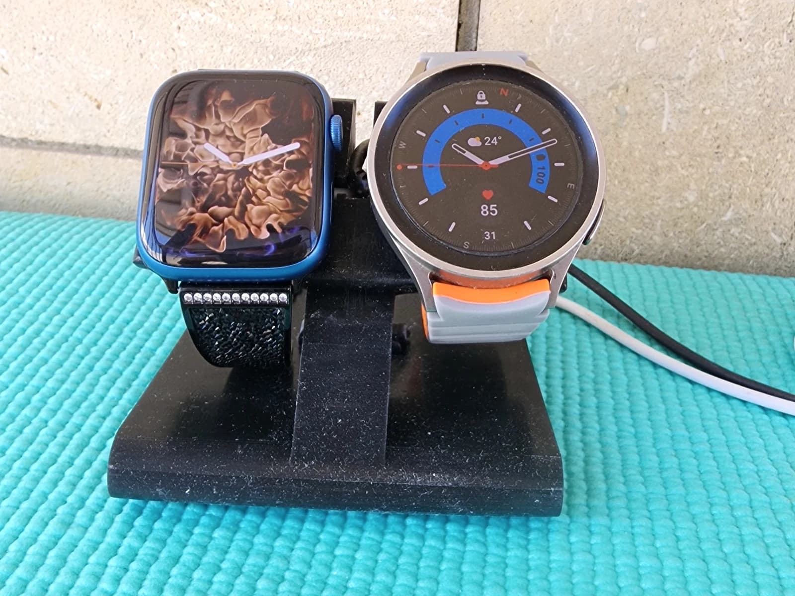 A black desktop stand holding two smartwatches on a turquoise surface.