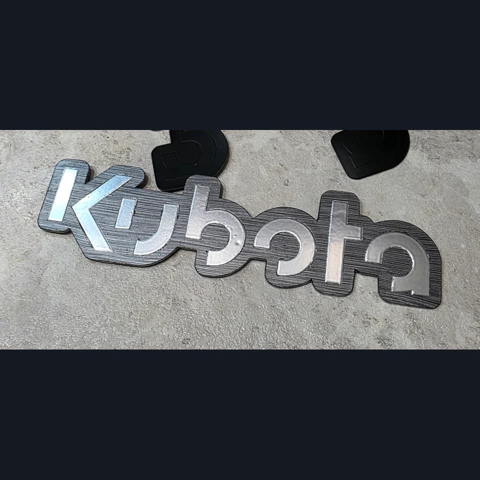 Silver-coloured Kubota lettering on a dark shaped badge, resting on a grey work surface.