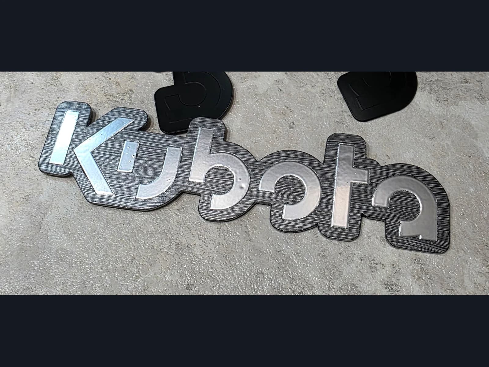 Silver-coloured Kubota lettering on a dark shaped badge, resting on a grey work surface.