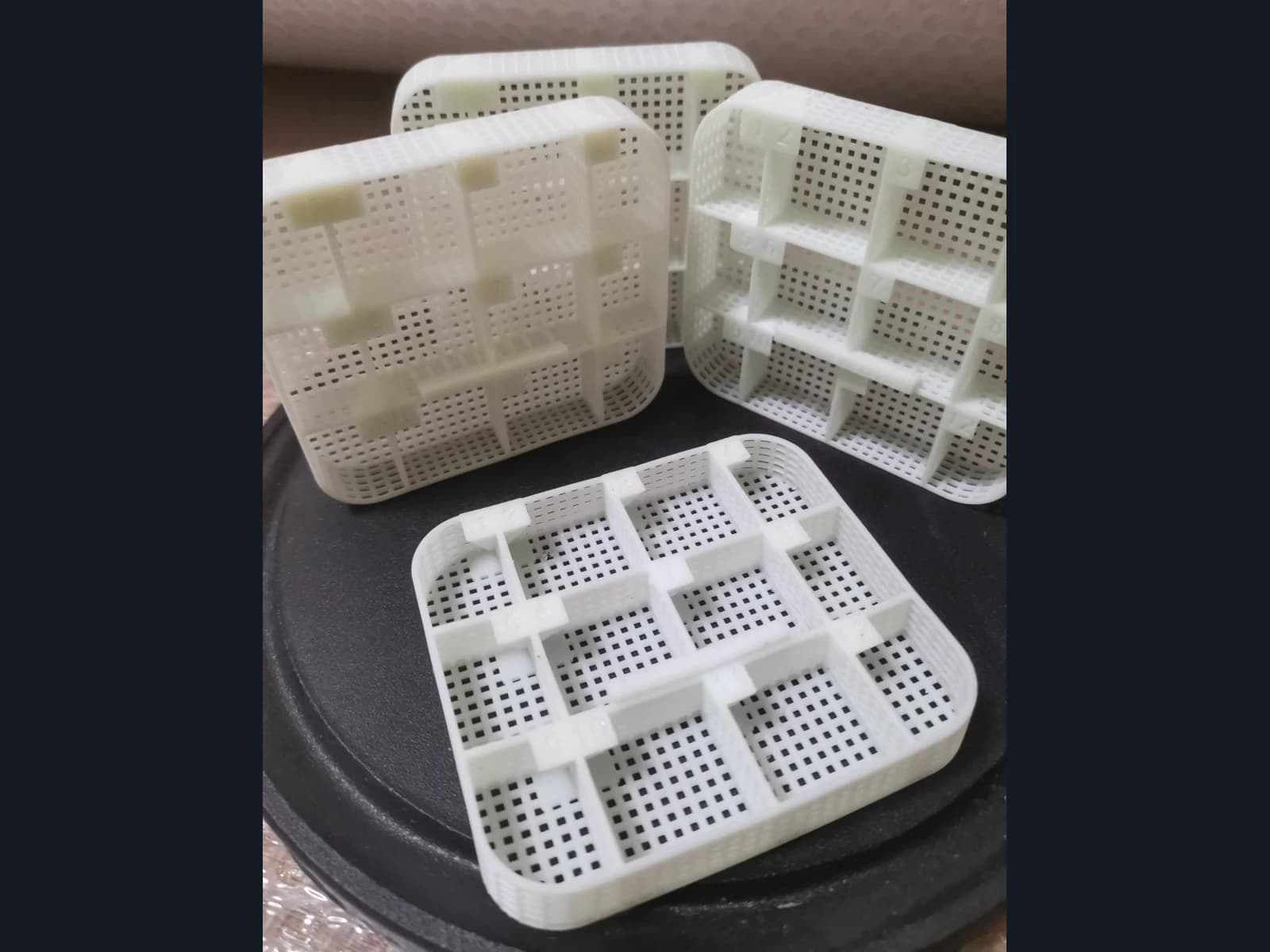 White perforated trays with internal dividers and tabs along their upper edges on a dark circular stand.