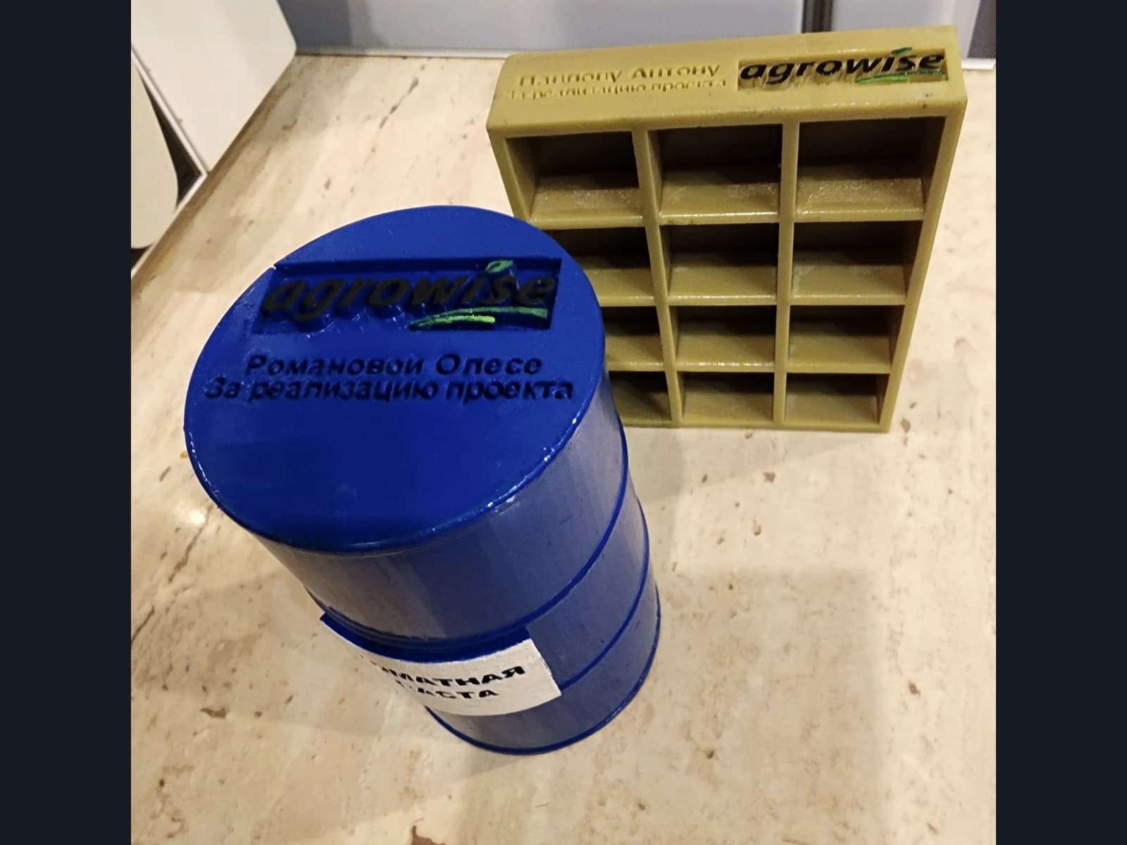 A blue barrel-shaped souvenir and a gold-coloured rectangular piece with compartments, both with Agrowise logos and lettering.