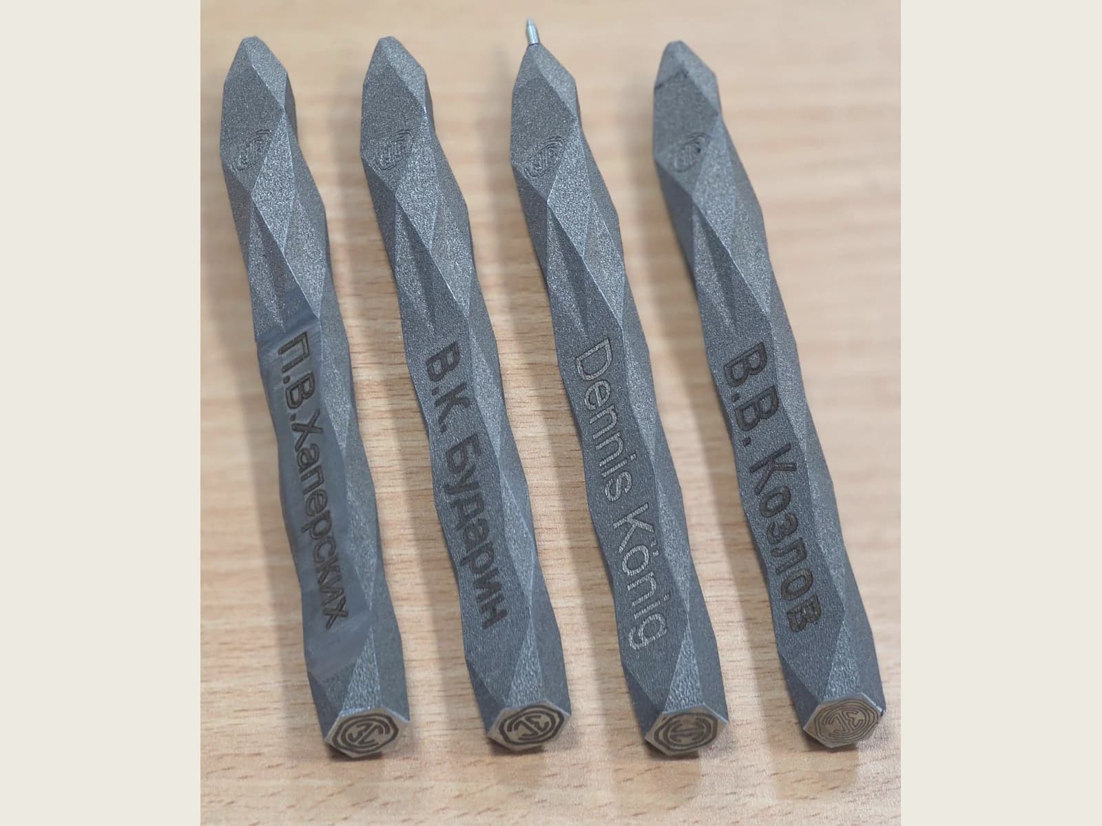 Four grey personalised pens with faceted bodies and circular emblems on their end faces, on a wooden surface.