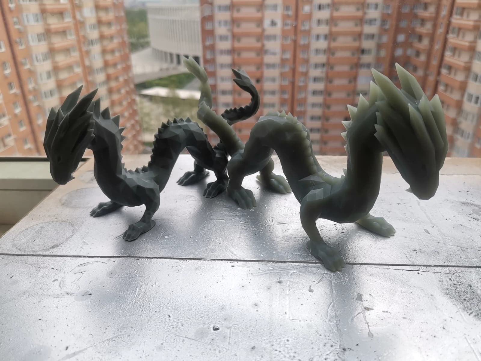 Two grey dragon figurines with faceted surfaces, curved necks and raised tails on a windowsill.