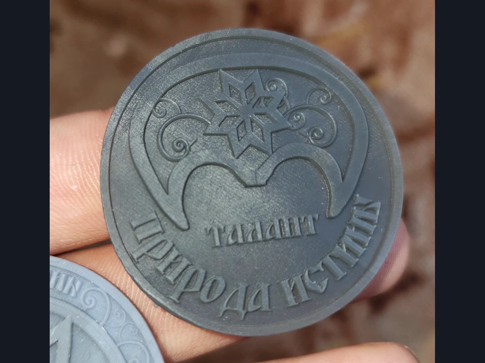 A grey relief token held in a hand, with an ornamental motif and the Russian words “Talent” and “Nature of Truth”.