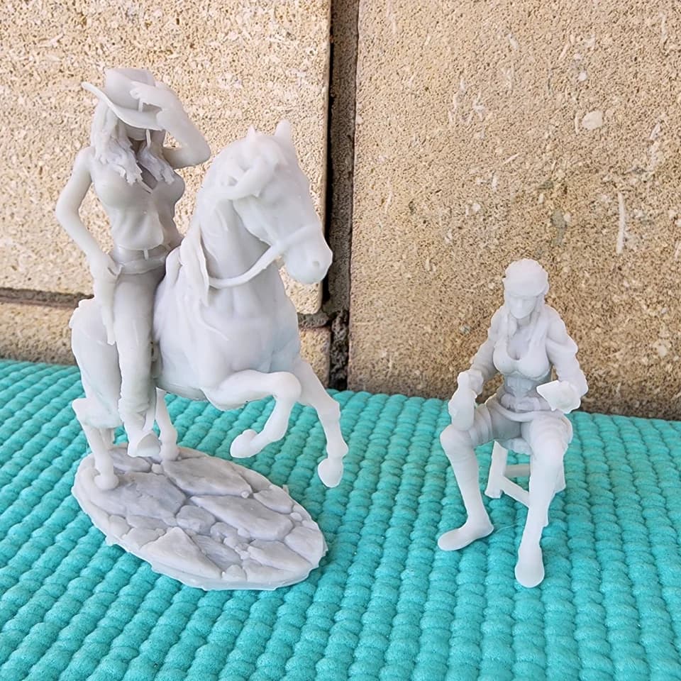 Two light-coloured figurines: a cowgirl riding a rearing horse and a seated woman in a hat, on a turquoise surface.