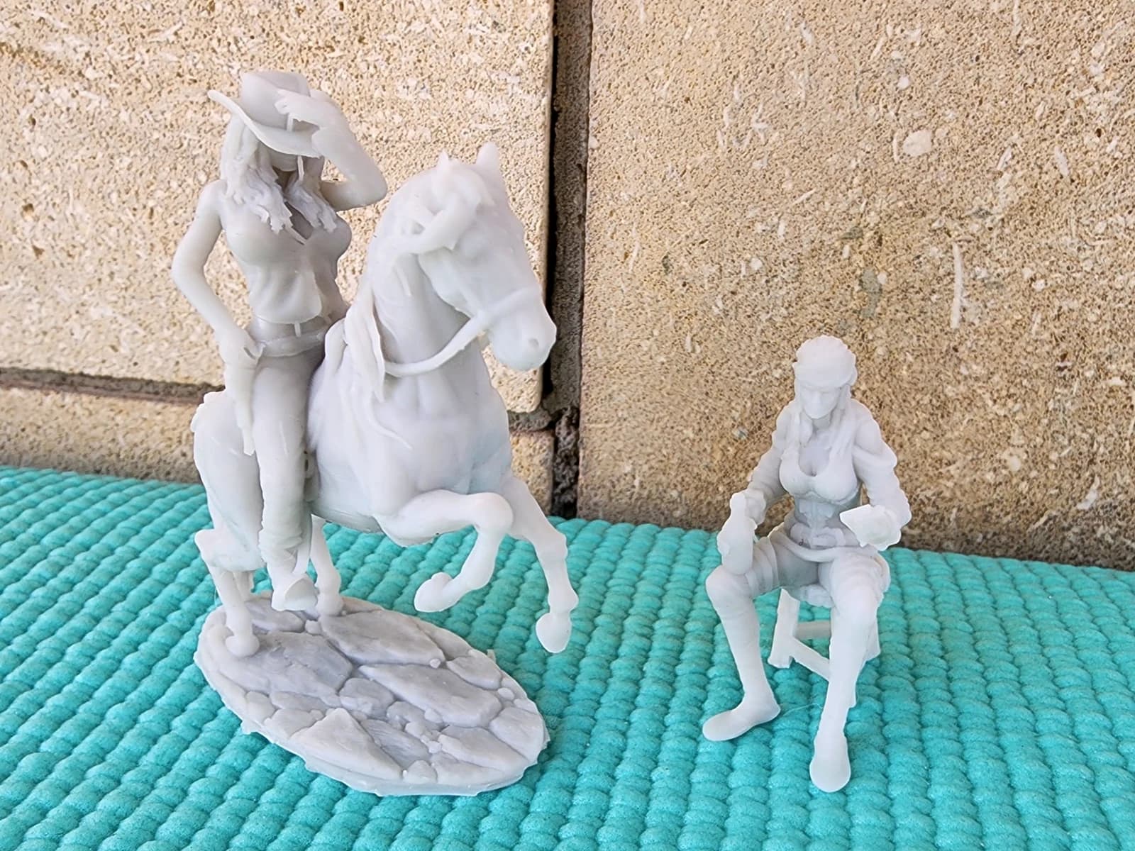 Two light-coloured figurines: a cowgirl riding a rearing horse and a seated woman in a hat, on a turquoise surface.