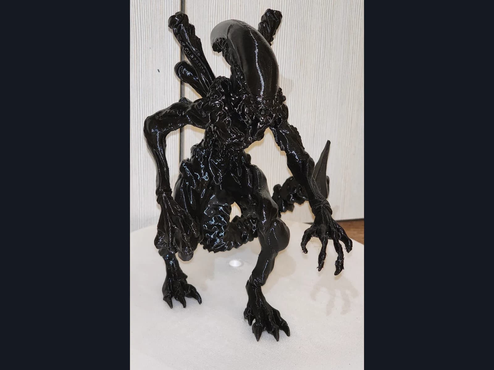 A glossy black Xenomorph figurine with an elongated head, tail and claws on a light platform.