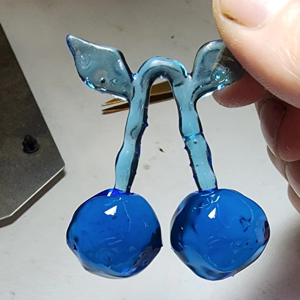 A translucent blue pendant shaped like two cherries with stems and leaves, held in a hand.