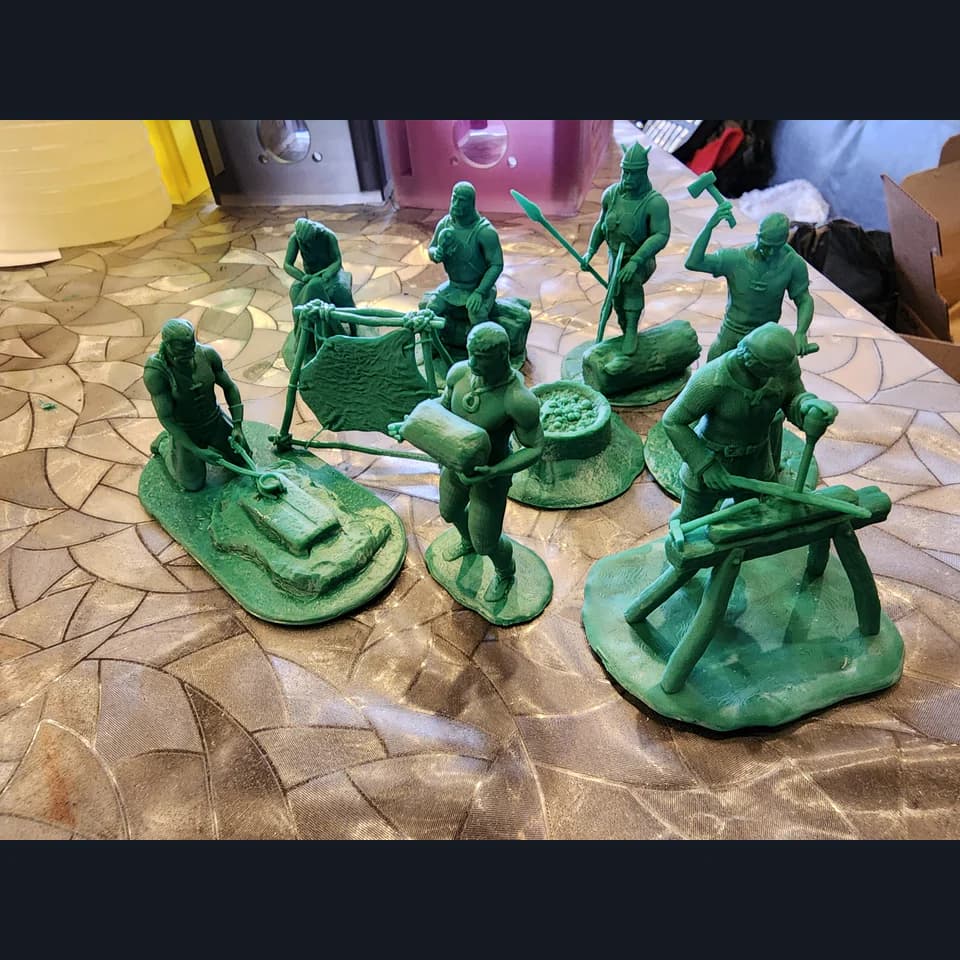 Green miniatures of people with tools and workpieces, including figures at an anvil and a workbench, on textured bases.