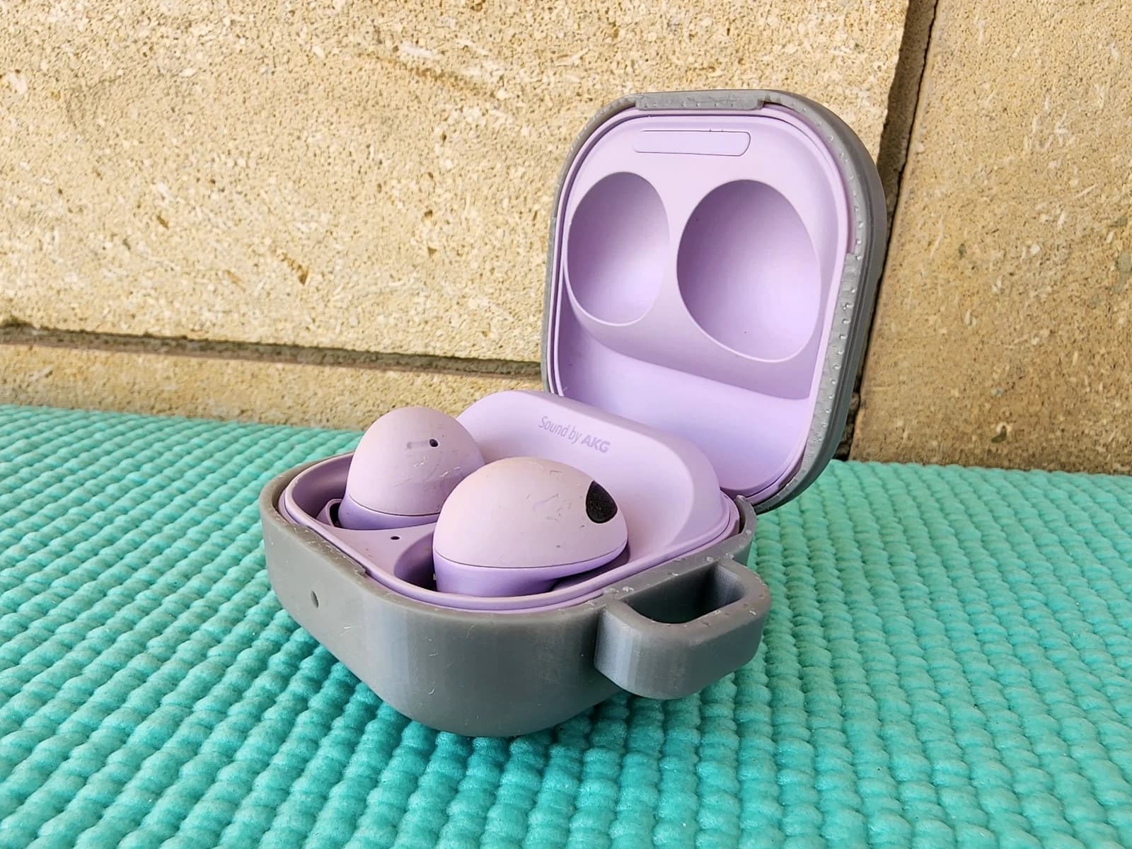 A grey cover with a side loop fitted around an open lilac wireless earbud case.