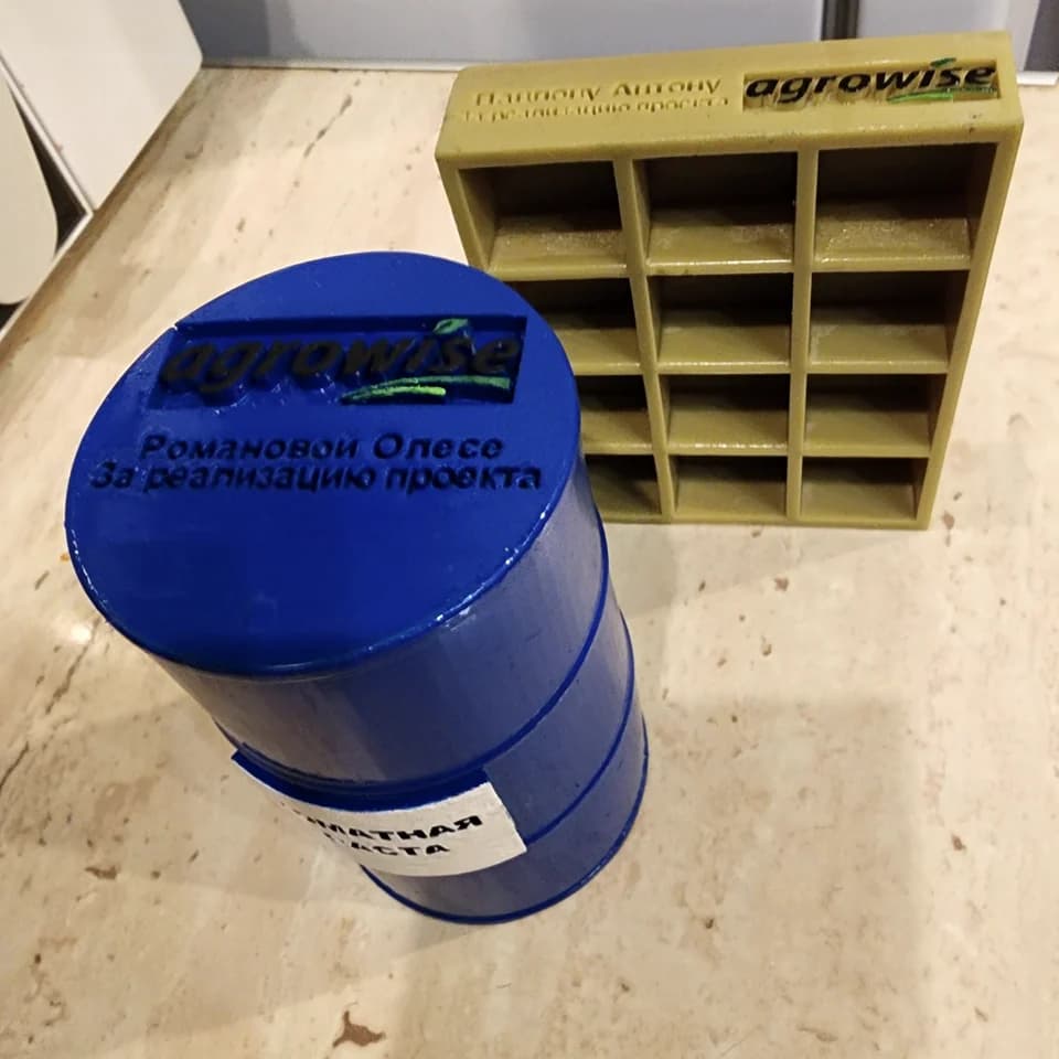 A blue barrel-shaped souvenir and a gold-coloured rectangular piece with compartments, both with Agrowise logos and lettering.