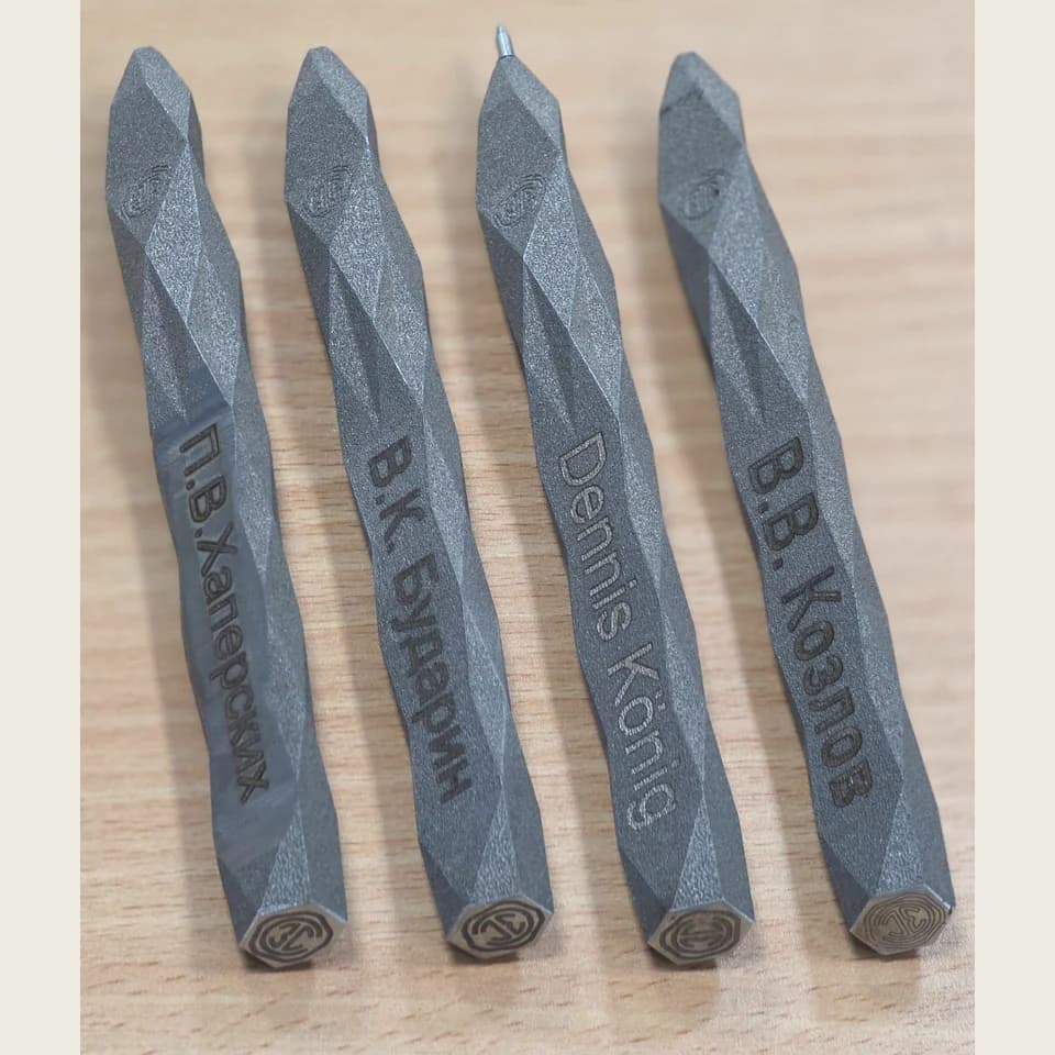 Four grey personalised pens with faceted bodies and circular emblems on their end faces, on a wooden surface.
