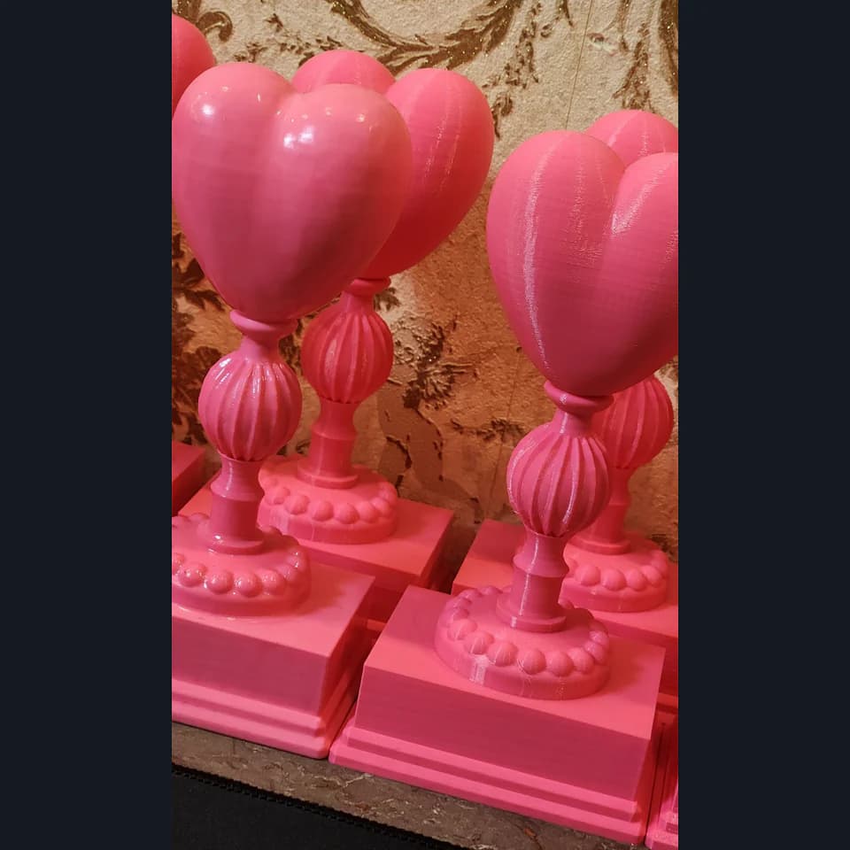 Pink decorative trophies with three-dimensional hearts, ribbed stems and stepped square bases, arranged next to each other.