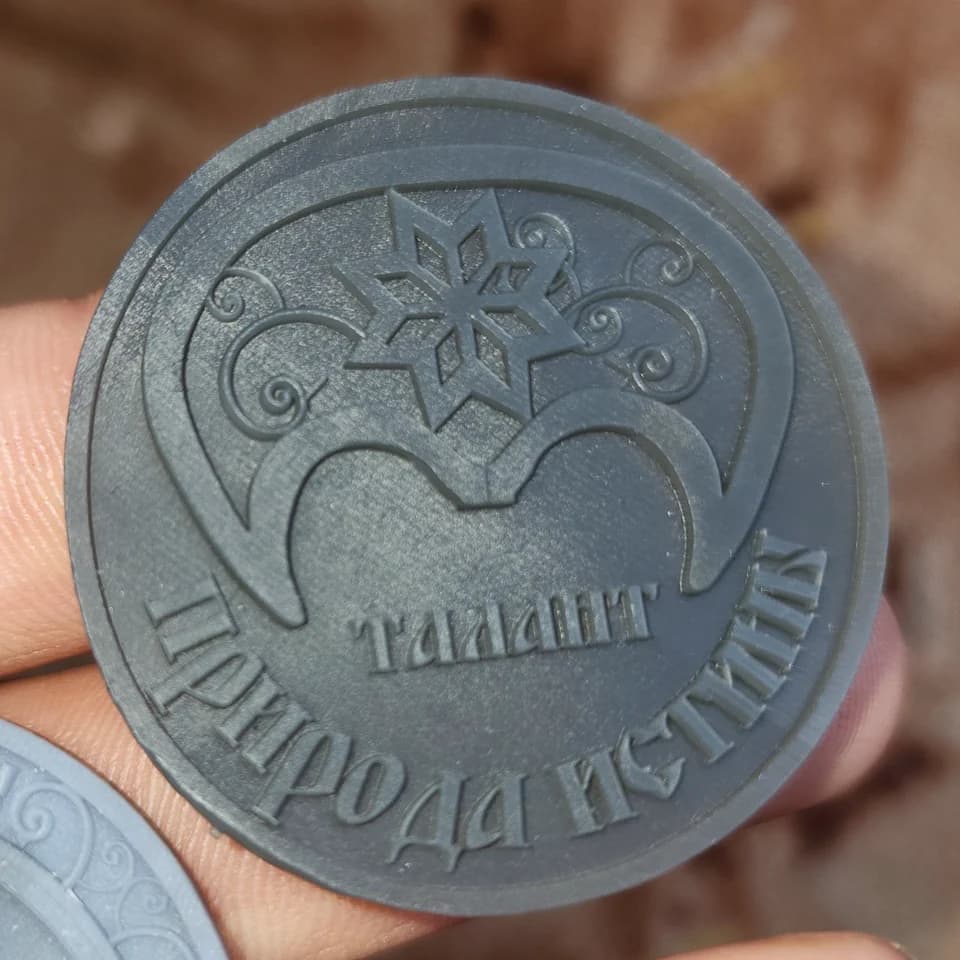 A grey relief token held in a hand, with an ornamental motif and the Russian words “Talent” and “Nature of Truth”.