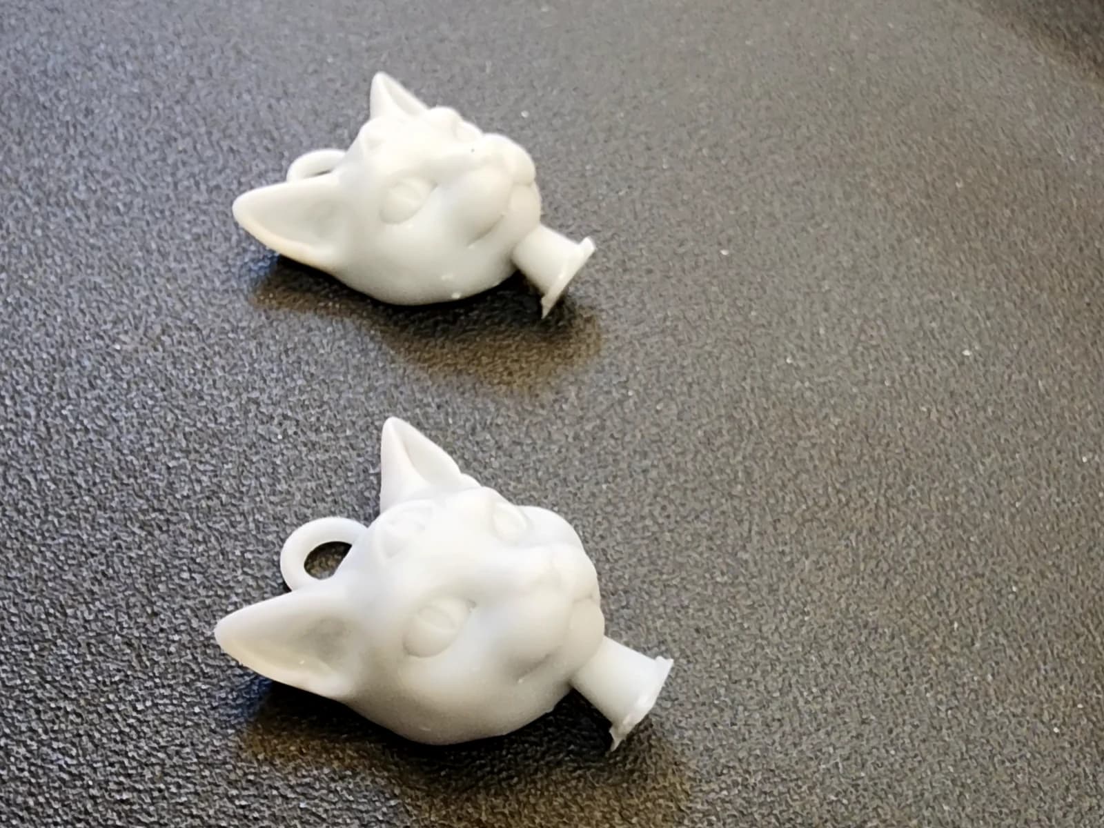 Two light-coloured three-eyed cat-head pendants with hanging loops on a dark textured surface.