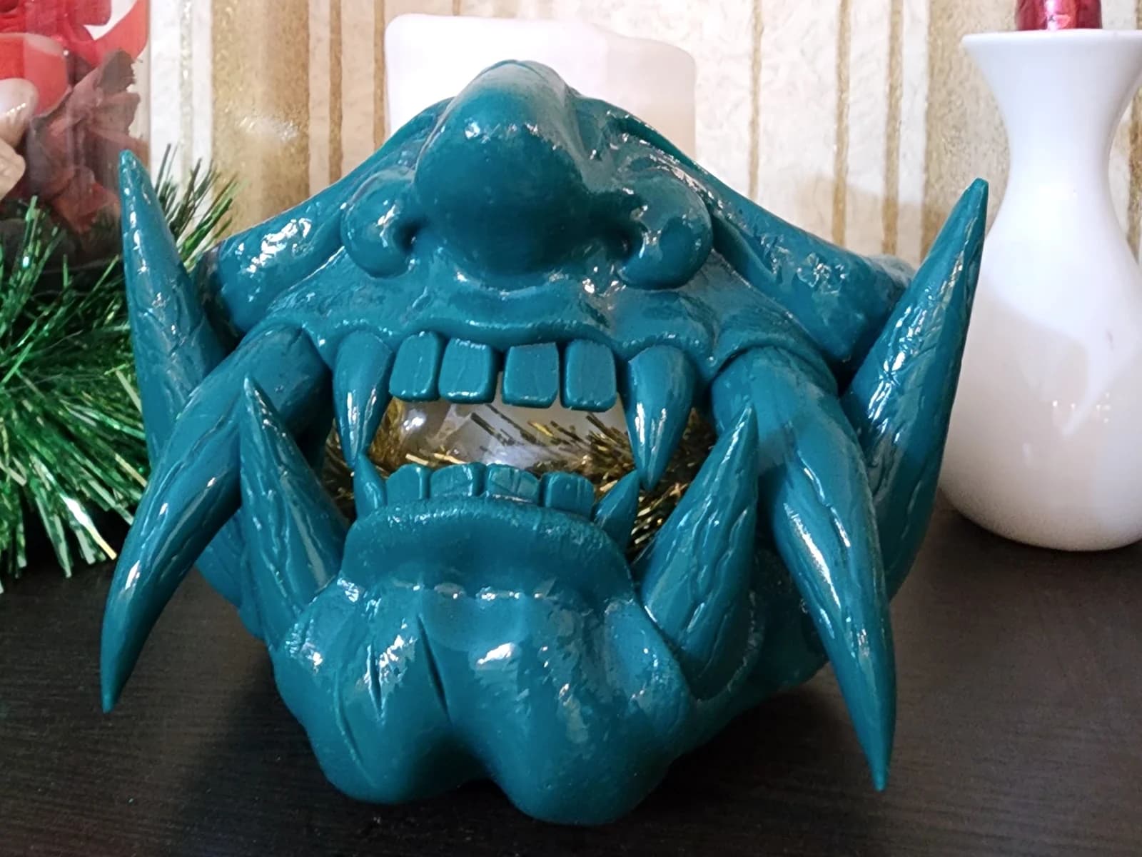 A turquoise orc mask with large tusks, teeth and pronounced facial relief on a dark shelf.